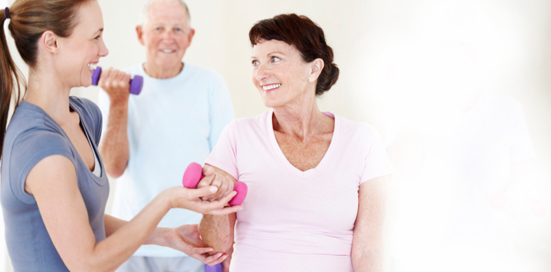 An elderly woman lifting a dumbbell while being helped be her instructor